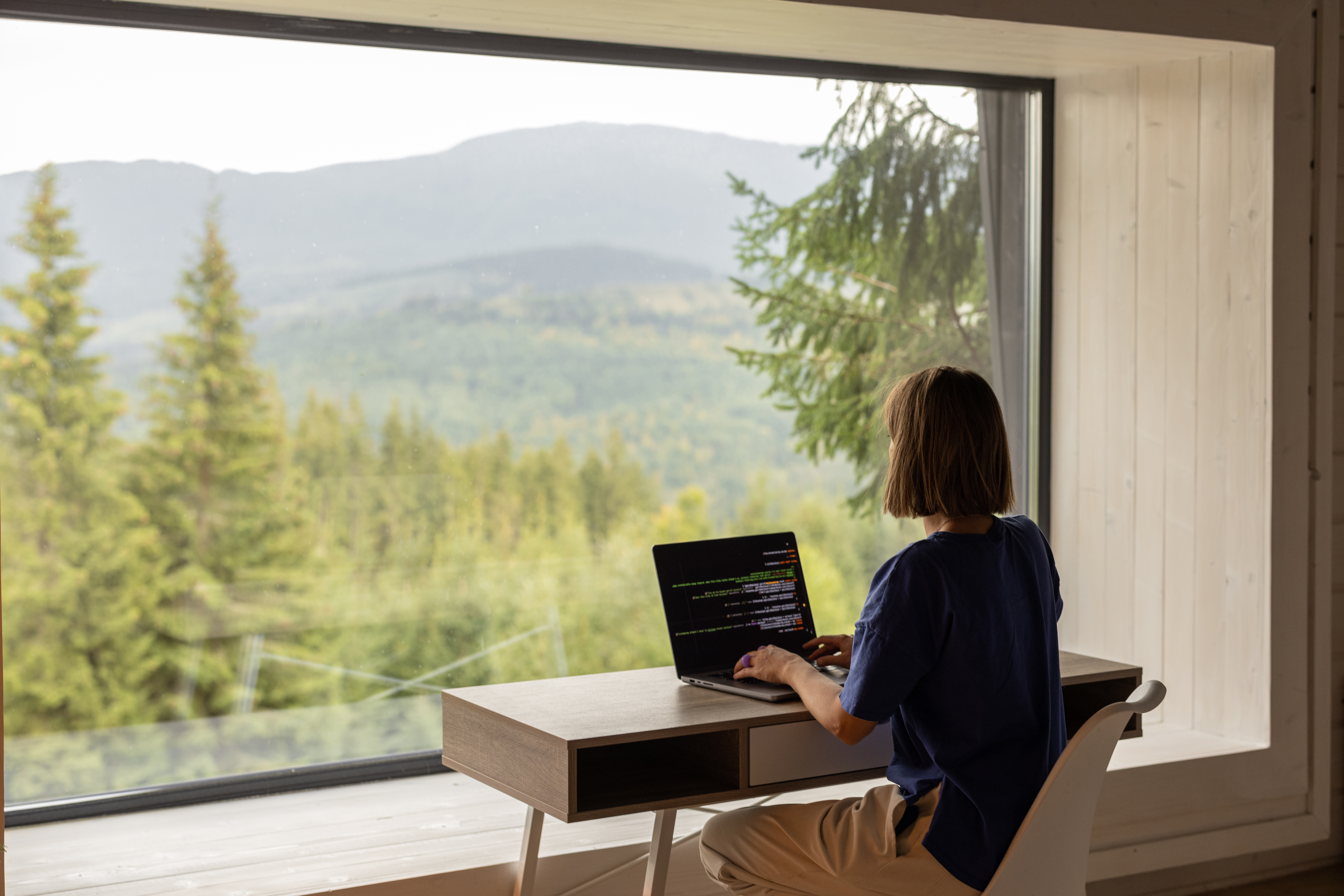En kvinne sitter ved en pult og skriver på en PC foran et stort vindu som viser en utsikt over en grønn barskog.