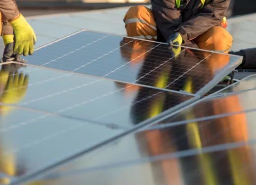 To par hender med arbeidshansker installerer nye, blanke solcellepaneler på bakken.