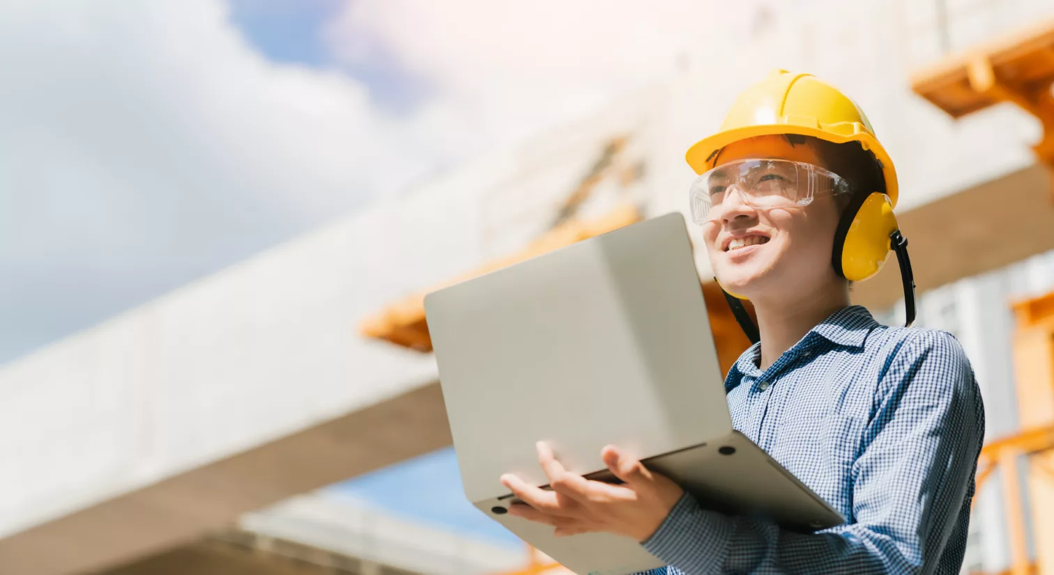 Person med gul hjelm, vernebriller og gule hørselsvern holder en PC i hendene og ser opp med et smil om munnen. I bakgrunnen er det en byggeplass med betongblokker under konstruksjon.