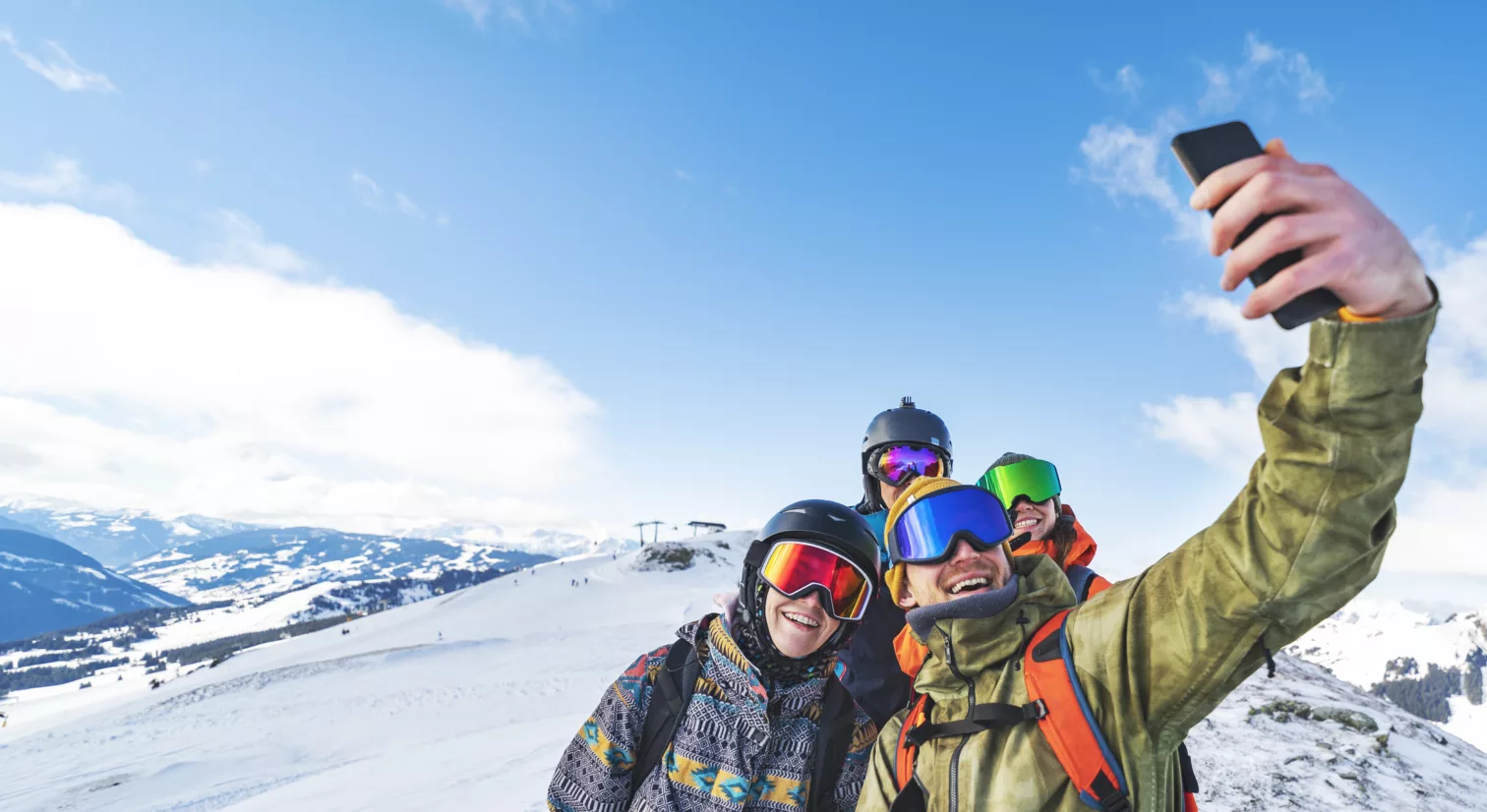 En gruppe på fire unge mennesker med vinter-antrekk, ryggsekker og slalombriller, står på toppen av et snødekt fjell. Det er blå himmel og skyer og snø i bakgrunnen. Den fremste personen i gruppen holder en telefon i hånden og tar en selfie av alle.