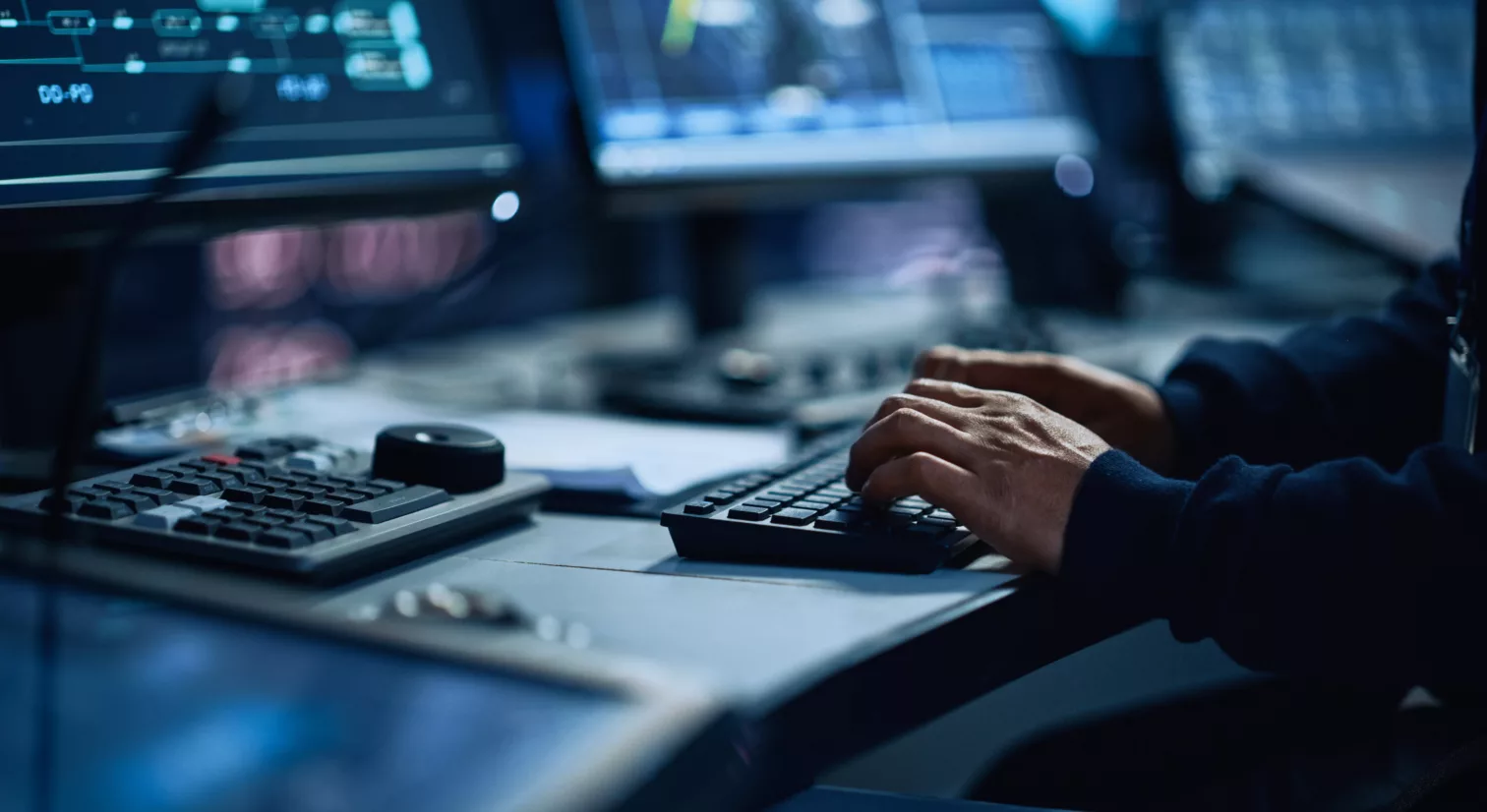 Nærbilde av hender som klikker på et tastatur under flere dataskjermer på en pult. Det er mørkt og det lyser blått fra skjermene.