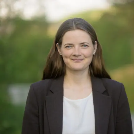 Portrett av smilende kvinne med mørkebrunt langt hår, i ført en hvit bluse med mørk blazer, bakgrunnen viser blå himmel og grønn natur med trær
