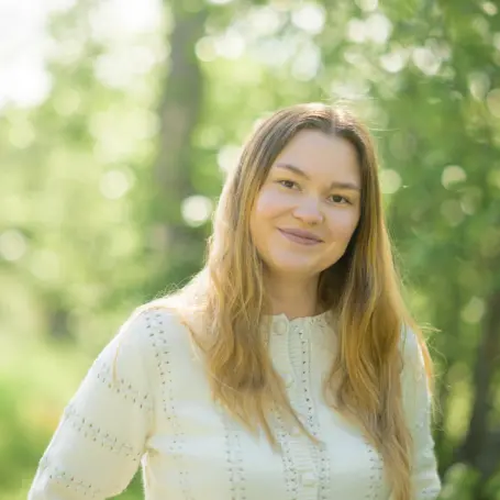 Bilde av smilende kvinne med lyst langt hår i ført en hvit genser, bakgrunner er uskarp men viser trær og sol