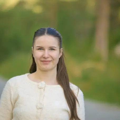 Portrett av kvinne med langt mørkt hår, iført en hvit cardigan som er knappet igjen, bakgrunnen er uskarp, men viser en grusvei omringet av grønne trær.