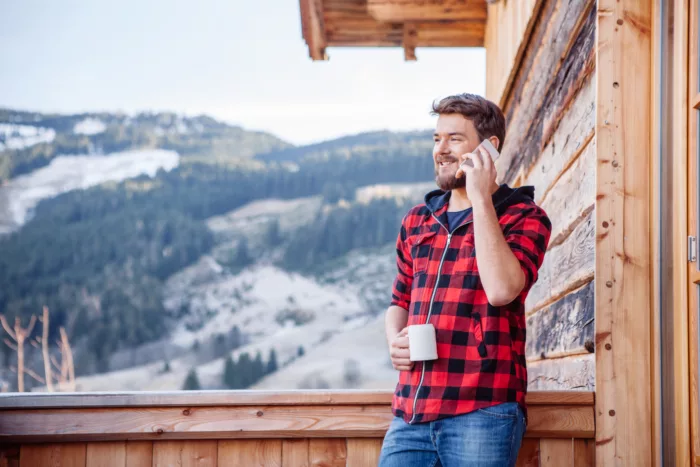 En mann med rutete og rød skjorte står utenfor en trehytte med en kaffekopp i hånden. Han snakker i telefonen og det er et nordisk fjellandskap i bakgrunnen.